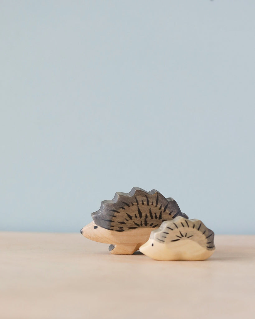Two Handmade Holzwald Hedgehog Baby figurines on a smooth surface against a plain blue background, with the smaller figurine in front and a larger one slightly behind it.