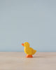 A small, yellow, high-quality Handmade Holzwald Duckling figurine stands on a solid surface against a soft blue background. The duck is profiled to the left with a simple, cartoonish design.