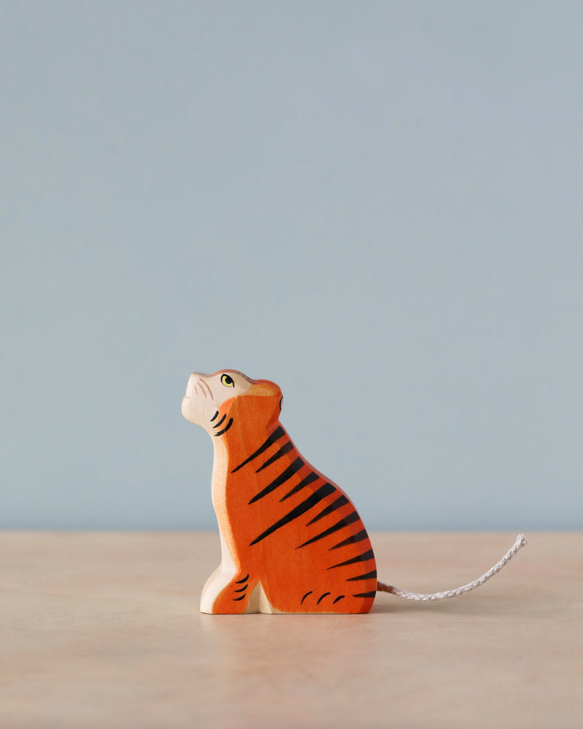 A Handmade Holzwald Sitting Tiger with a whimsical design, painted in bright orange with black stripes, sitting upright and looking upwards. The tiger has a white underbelly and a small tail with a string attached.