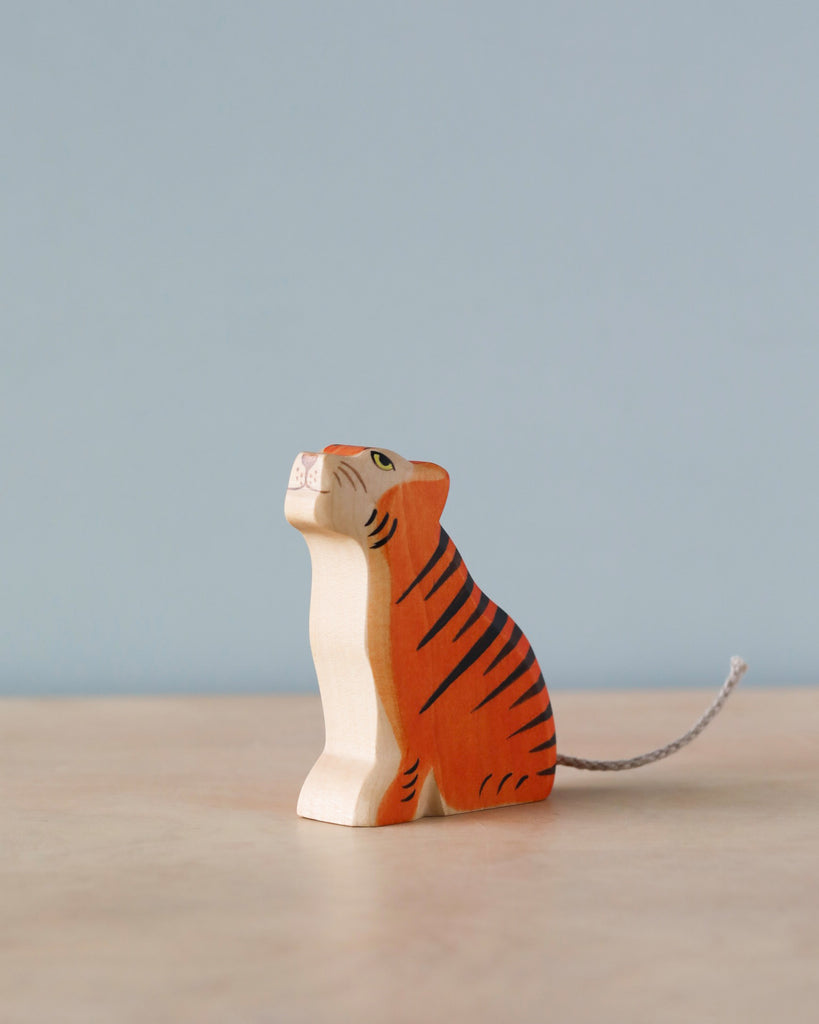 A sustainable Handmade Holzwald Sitting Tiger figurine with black stripes, standing against a soft blue background. The tiger is depicted sitting and looking upwards, with a finely detailed face and a small, curved tail.