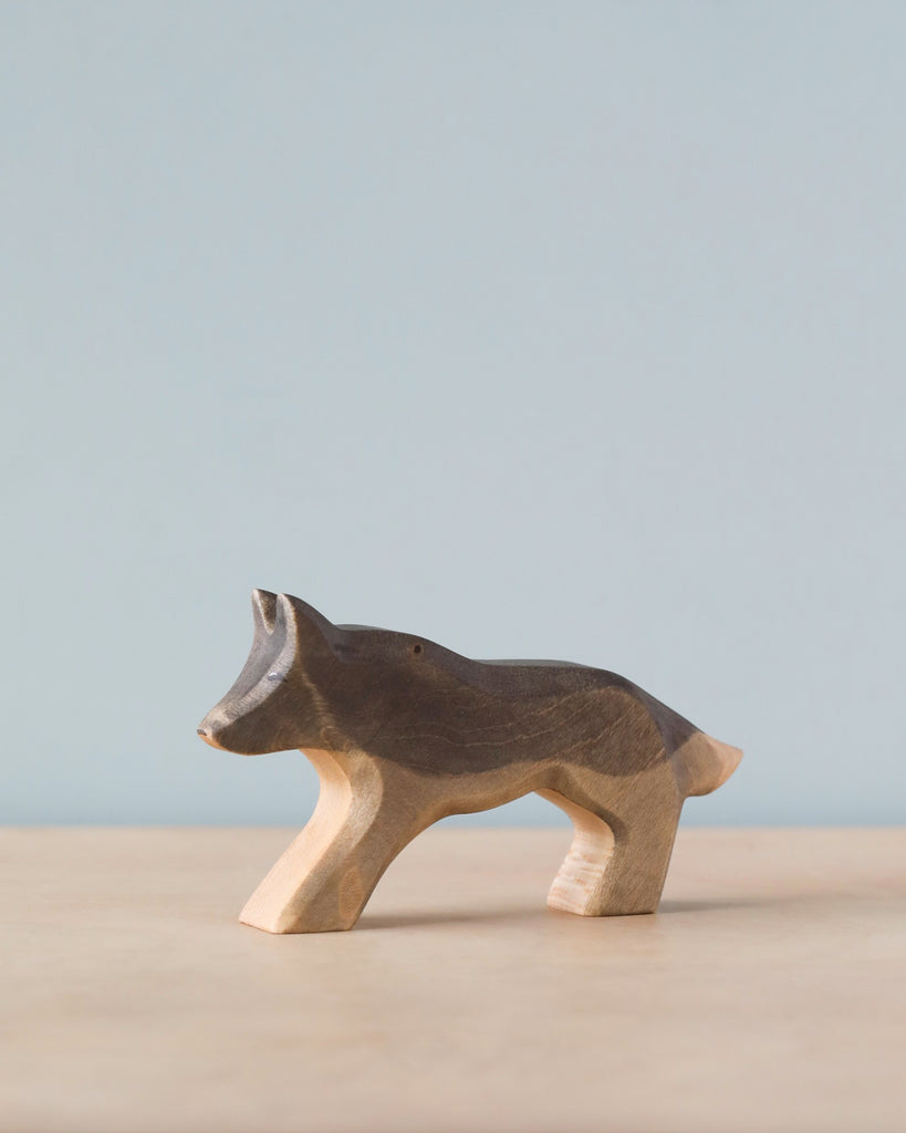 A Handmade Holzwald Wolf figurine against a soft blue background stands on a light brown surface. This high-quality wooden toy is carved in a simplistic style with smooth lines and visible wood grain.