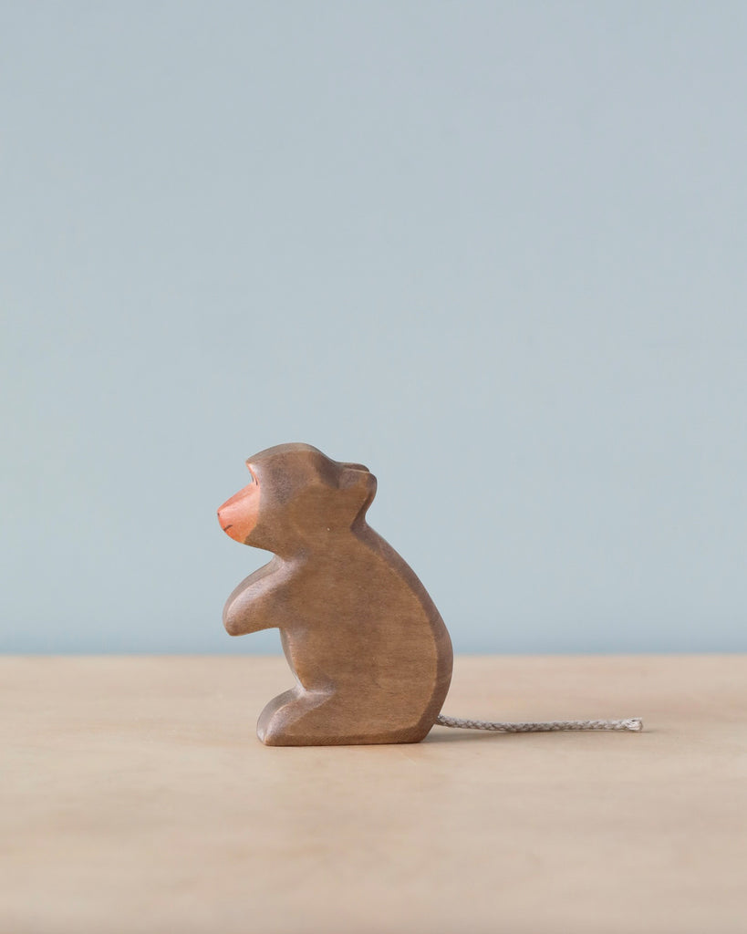 A Handmade Holzwald Baboon wooden mouse figurine with a pink nose and a thin rope tail, standing on a flat surface against a soft blue background.