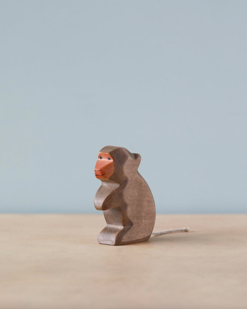 A Handmade Holzwald Baboon with a prominent orange face, sitting against a soft blue background. This sustainable toy is textured and appears handcrafted, with its tail curling to the side.