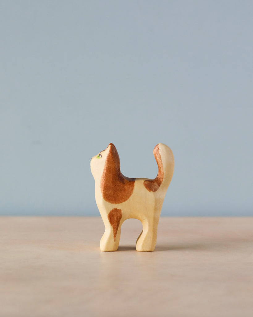 A Handmade Holzwald Cat figurine, crafted from high-quality wood, with a light tan body and brown patches, standing upright against a plain blue background.