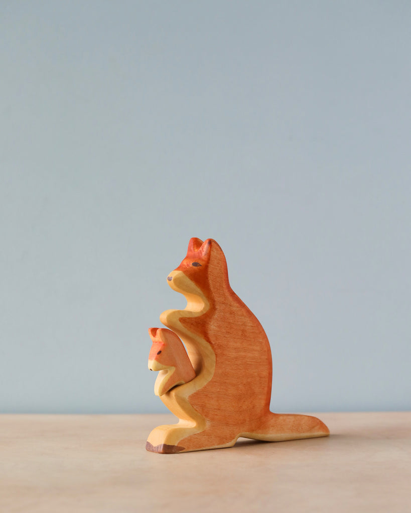 A Handmade Holzwald Kangaroo With Baby figurine stands on a flat surface against a soft blue background. The figurine, representing sustainable toys, is intricately carved, depicting the kangaroo with her baby in a sitting pose.