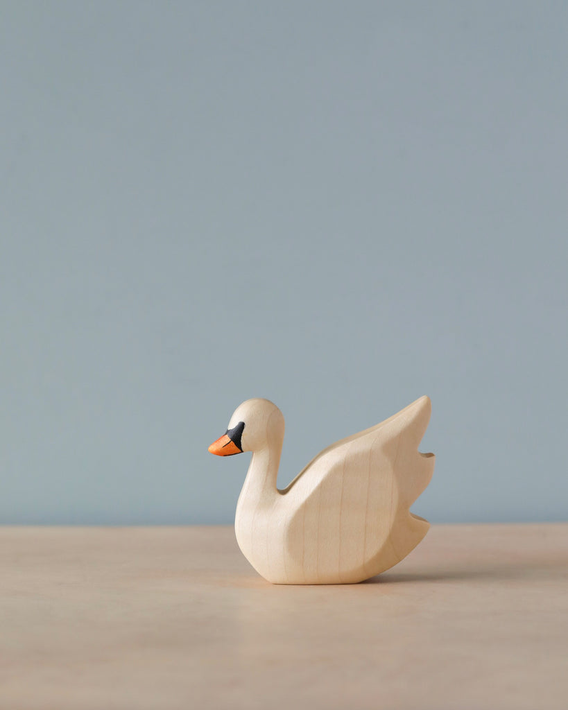 A simplistic Handmade Holzwald Swan figurine, crafted with natural dyes, stands on a light beige surface against a soft blue background. The swan has a gracefully curved neck and minimalistic details.