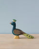 A Handmade Holzwald Peacock figurine stands against a soft blue background, featuring intricate details on its plumage and a proud, upright posture. This piece is one of the sustainable toys in our collection.