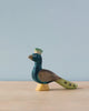 A Handmade Holzwald Peacock, painted with high-quality oil in shades of teal and navy, stands on a small yellow base against a plain, light blue background.