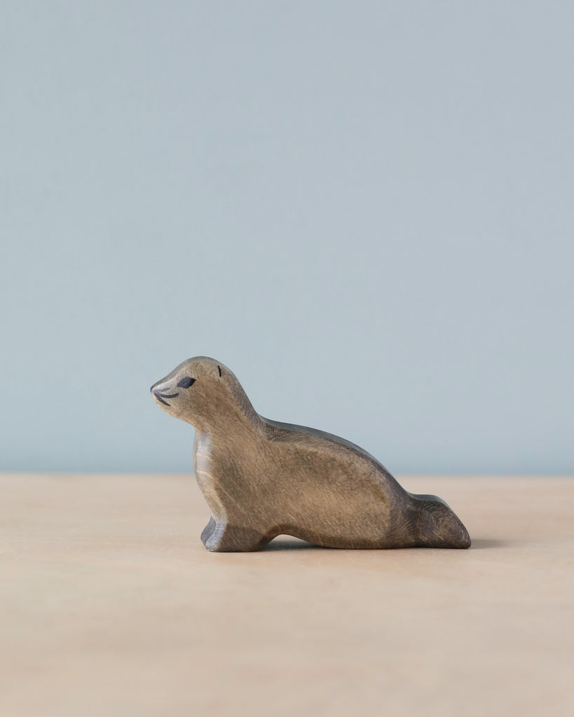 A Handmade Holzwald Baby Sea Lion carving resting on a flat surface against a soft blue background. The sea lion, crafted from high-quality wood, has a smooth, polished finish and a gentle, smiling expression.