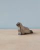 A small, intricately carved Handmade Holzwald Baby Sea Lion figurine, crafted from high-quality wood, positioned on a smooth wooden surface against a soft blue background.