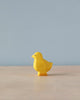 A small, bright yellow handmade Holzwald Baby Chick sits on a wooden surface against a pale blue background, depicting a simple and minimalist composition.