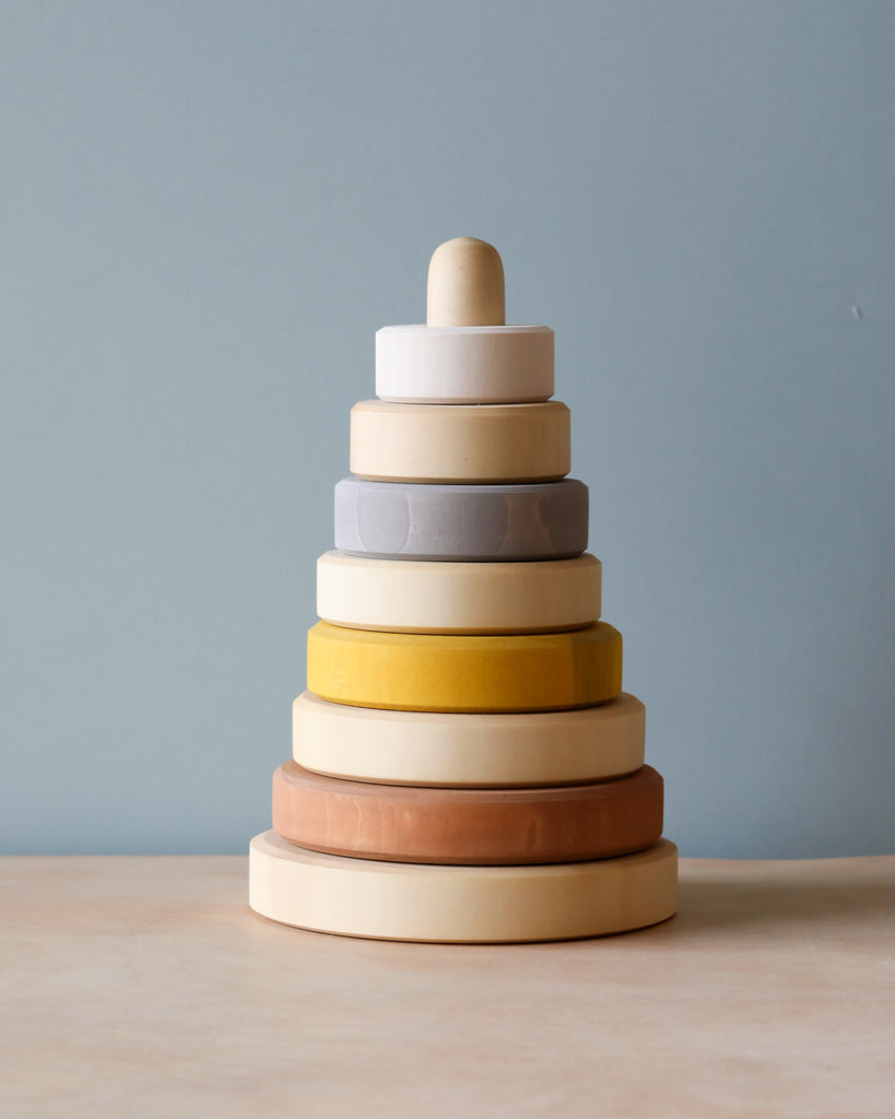 A Raduga Grez Handmade Pyramid Tower Stacker - Sand with rings in various sizes and muted colors, coated with non-toxic paint, topped with a round cap, placed on a wooden base against a light blue background.