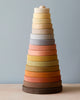 A Raduga Grez handmade large pyramid tower stacker featuring twelve rings in a gradient of pastel colors, painted with non-toxic paint, arranged in size order on a vertical column against a light blue background.