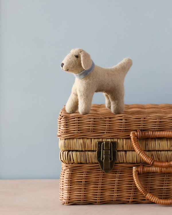 Sentence with product name: A Hand Felted Wool Golden Retriever Dog stands on a wicker box with a light blue background. The puppy is beige with a blue collar, and the box features a metal.