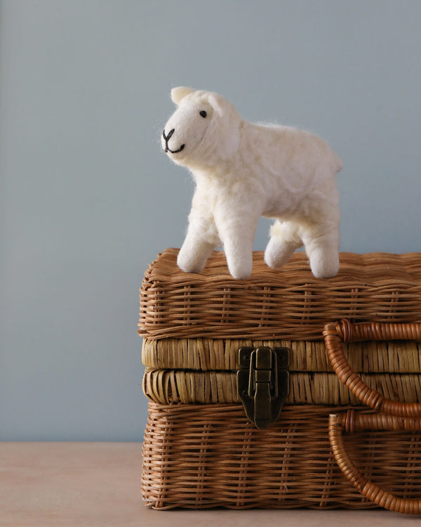 A handcrafted Hand Felted White Wool Sheep toy stands atop a woven wicker basket with a brass clasp, set against a soft blue background.