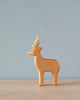 A small wooden Handmade Holzwald Male Deer figurine with detailed antlers stands against a soft blue background. The figurine is simplistic in style, showcasing smooth lines and a light, natural.