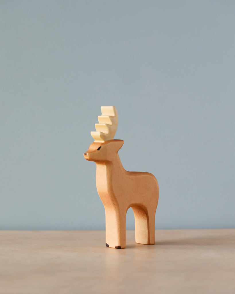 A Handmade Holzwald Male Deer figurine with an elongated, stylized antler, standing against a soft blue background on a light wooden surface.