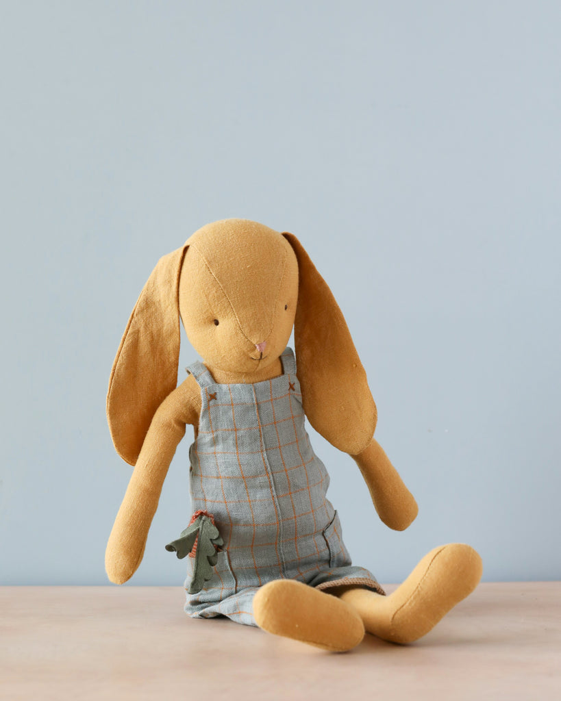 A Maileg Bunny Size 3, Dusty Yellow with long ears and a soft natural linen blue plaid dress, sitting against a plain blue background. The bunny appears well-loved, with a slight worn texture.