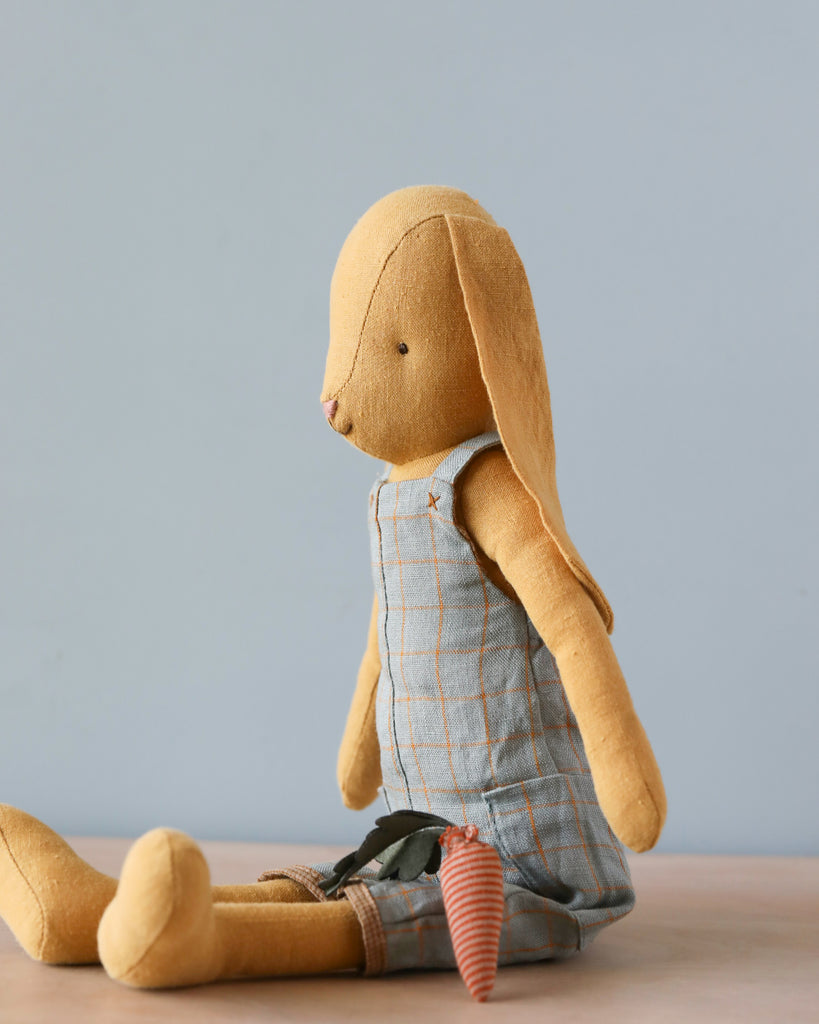 A soft natural linen Maileg Bunny Size 3, Dusty Yellow toy with stitched details, dressed in a plaid jumpsuit, sitting against a plain background. The toy is beige with flopped ears and an orange-striped limb.
