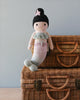 A Cuddle + Kind Luna The Mermaid doll with a black beanie and pink outfit, filled with hypoallergenic polyfill, sits on a wicker picnic basket against a neutral background.