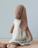 A Maileg Bunny Size 4, Brown - Knitted Dress with long, floppy ears is sitting upright. Dressed in a knitted light blue and beige checkered dress, the bunny is crafted from natural linen fabric and positioned against a light blue background on a wooden surface. Recommended age: all ages.