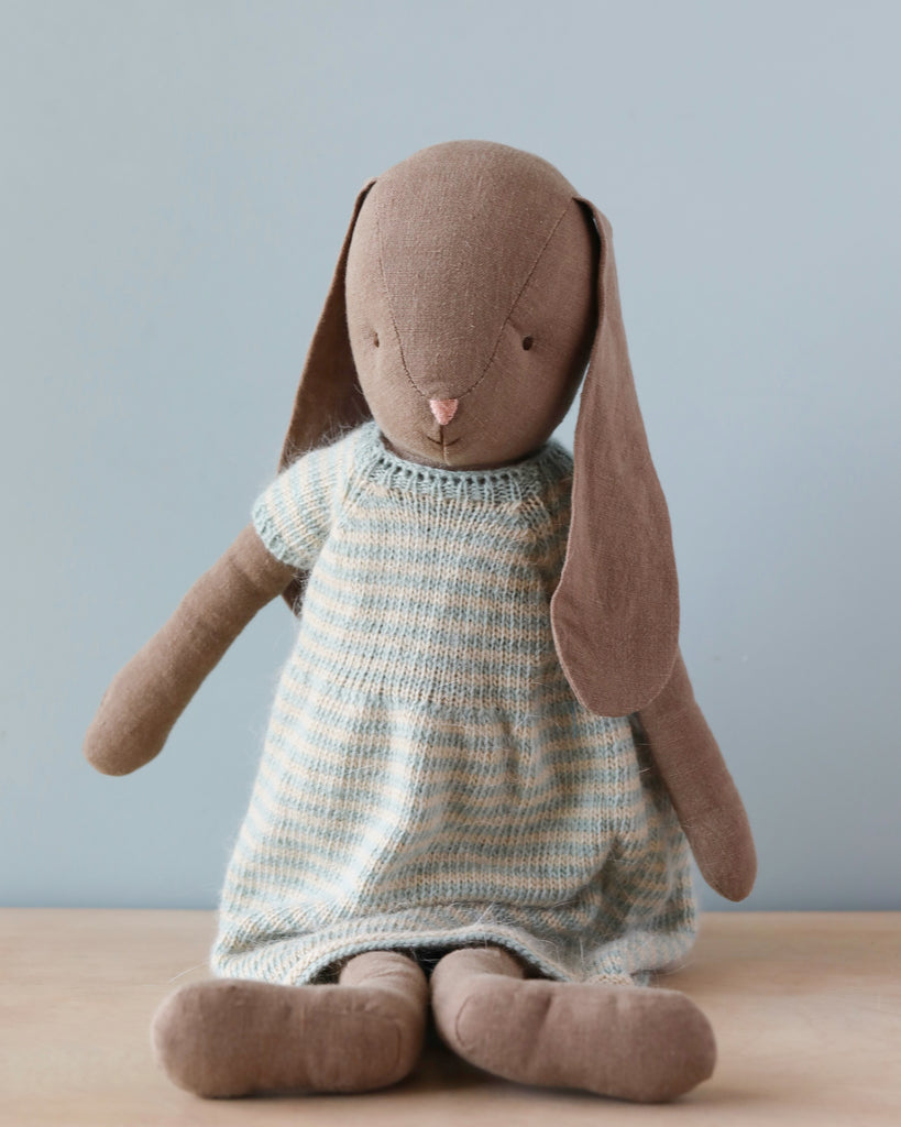 A Maileg Bunny Size 4, Brown - Knitted Dress sits upright against a light blue background. Made from natural linen fabric, it has long ears and wears a blue and white knitted dress with a checkered pattern. The bunny's features are minimal, with small black eyes and a pink nose. Recommended age: all ages.