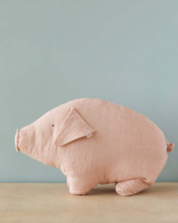 The Maileg Medium Polly Pork is a charming pig-shaped plush pillow featuring a simplistic, minimalist design with light pink fabric and subtle stitched details for the eyes and nostrils. Perfect for cuddles, this adorable pig rests on a wooden surface against a light blue backdrop.