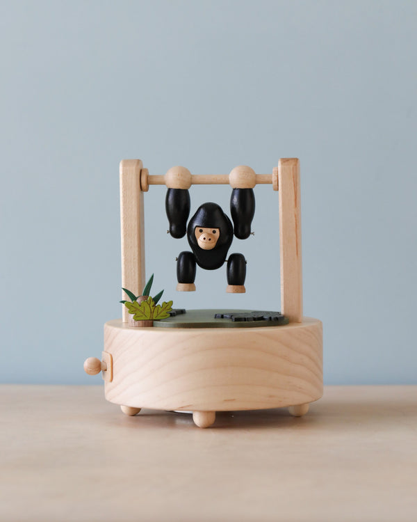 A Gorilla Music Box featuring a small monkey figure with black limbs and a neutral expression, swinging from a horizontal bar. The base includes a small green plant and is crafted from sustainably sourced wood.