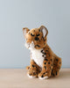 A sitting jaguar cub stuffed animal sits against a plain blue background, featuring an artisan hand-sewn design with a focused expression, prominent whiskers, and spotted orange fur.