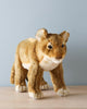 A realistic Standing Lion Cub Stuffed Animal, artisan crafted, standing on a wooden surface against a soft blue background. The toy displays detailed features such as fur texture and facial markings.