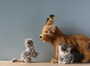 Three Pallas Kitten Stuffed Animals, including a grey kitten, a terrier dog, and a small duck sitting on the dog's head, are arranged on a wooden surface against a pale blue background.