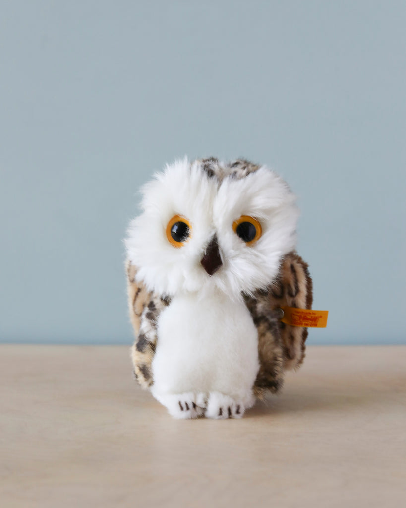A Steiff, Wittie Owl plush toy with large, striking yellow eyes and soft, speckled brown and white feathers, standing against a plain blue background. This toy is crafted from cuddly soft woven fur.