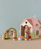 The Rosewood Cottage is a pastel-colored wooden toy house with floral designs and heart windows, reminiscent of a woodland cottage. Small wooden figures in mushroom-shaped hats and colorful outfits gather around it, along with a tiny flower table and arch.