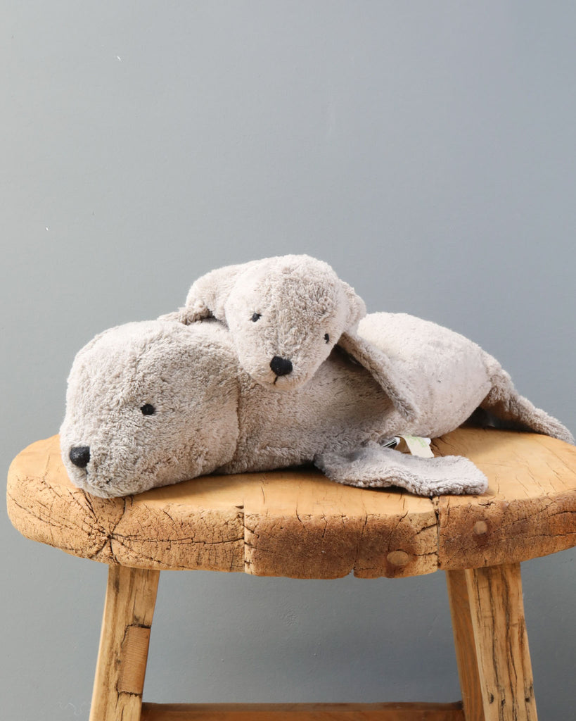 Two Senger Naturwelt Cuddly Animal - Gray Seal toys lying on a rustic wooden stool against a soft gray background. The toys appear well-loved, with one resting its head on the other's back.