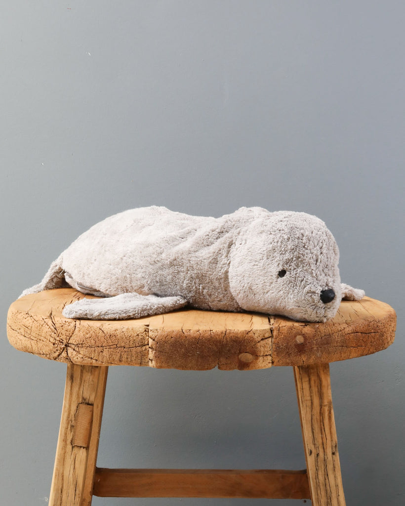A Senger Naturwelt Cuddly Animal - Gray Seal lying on a rustic wooden stool against a grey wall. The seal is grey and looks soft and fluffy, capturing a serene and cozy appearance.