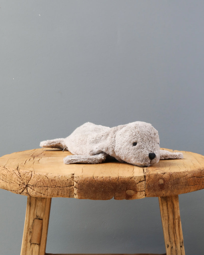 A Senger Naturwelt Cuddly Animal - Gray Seal lying on a rustic wooden stool against a plain grey backdrop. The seal is light grey with a simple, cute face.