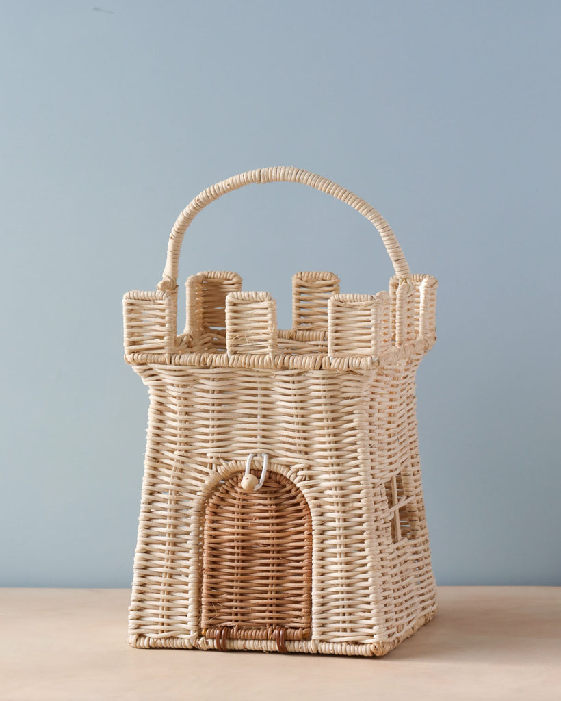 An Olli Ella Rattan Castle Bag shaped like a magical castle with a handle, featuring details such as windows and a doorway, set against a plain light blue background.