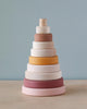 A handmade Wooden Pyramid Stacker - Pink with rings in various sizes and soft pastel colors including pink, yellow, and cream, arranged in ascending order, set against a light blue background.