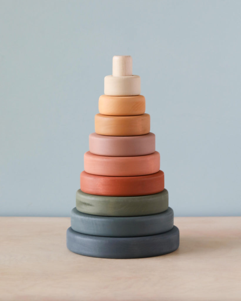 Sentence with product name: A Wooden Pyramid Stacker - Green & Mustard coated with non-toxic paint, featuring rings in various muted colors including light blue, green, beige, and different shades of orange, arranged in order from largest at the bottom to