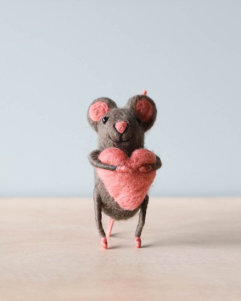 A small hand-felted grey mouse holding a pink heart in its arms. 