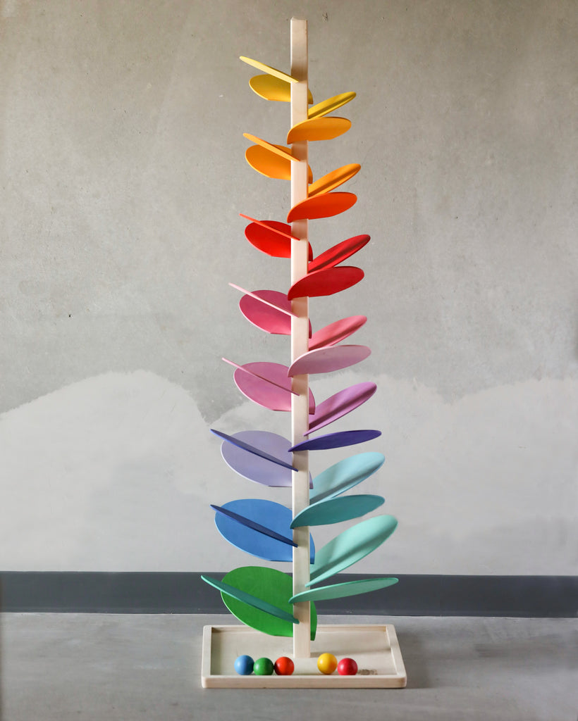 A colorful kinetic sculpture, dubbed the "Giant Magic Wood Marble Tree," features a series of horizontal, multicolored blades arranged along a vertical pole, set against a gray background, with wooden marbles at the.