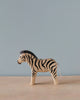 A Handmade Holzwald Zebra figurine stands on a plain surface against a pastel blue background, showcasing detailed black stripes and a small tail.
