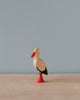 A Handmade Holzwald Stork figurine with a red beak and legs, and a black and white body, standing against a pale blue background.