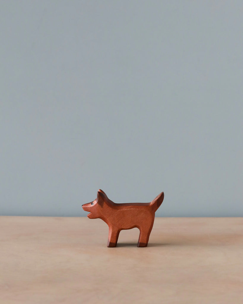 A small, high-quality Handmade Holzwald Brown Dog figurine stands upright against a plain blue-gray background. The figurine is simplistic, with minimal detailing on its body.
