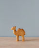 A small, simple Handmade Holzwald Baby Camel figurine stands on a smooth surface against a plain, light blue background. This high-quality figurine is crafted with minimalistic details, emphasizing the shape of the camel.