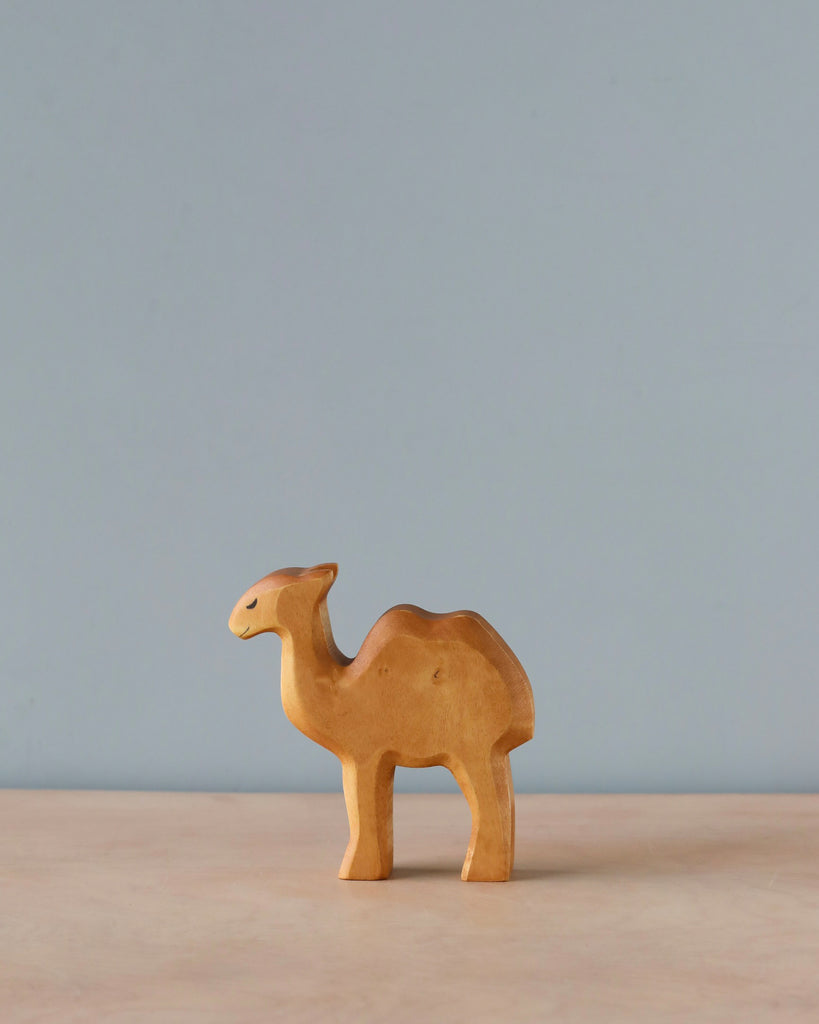 A small, simple Handmade Holzwald Baby Camel figurine stands on a smooth surface against a plain, light blue background. This high-quality figurine is crafted with minimalistic details, emphasizing the shape of the camel.