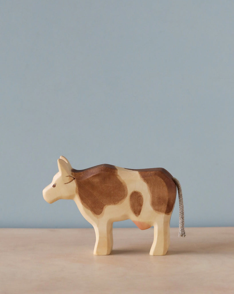 A Handmade Holzwald Cow with brown spots and a gray tail on a light blue background.