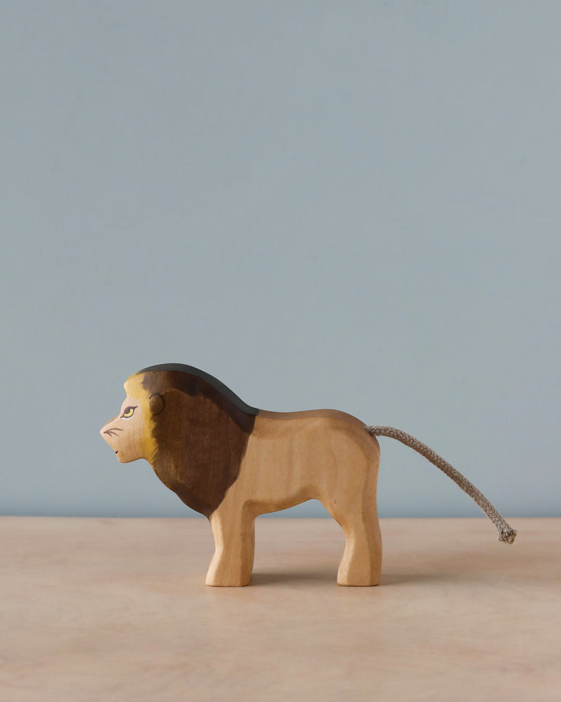 A Handmade Holzwald Lion figurine, crafted as part of a sustainable toys collection, stands against a soft blue backdrop. The lion has a detailed mane and tail, with its body carved to show muscle definition.