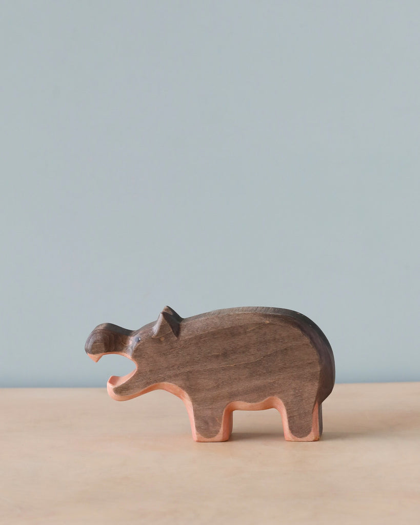 A Handmade Holzwald Hippo figurine on a smooth surface with a pale blue background. The figurine, representing sustainable toys, is crafted with visible details such as eyes and ears and has a natural wood