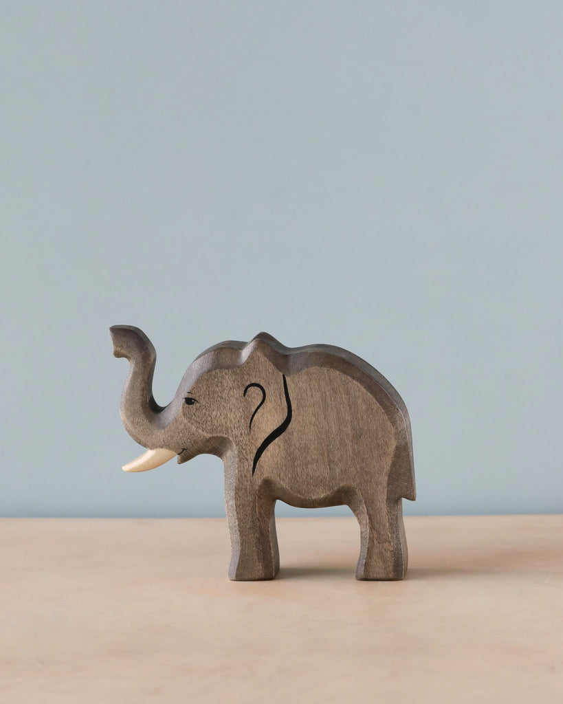 A Handmade Holzwald Elephant - Trunk Raised sculpture with visible ivory tusks, standing against a plain blue background with a light brown floor.